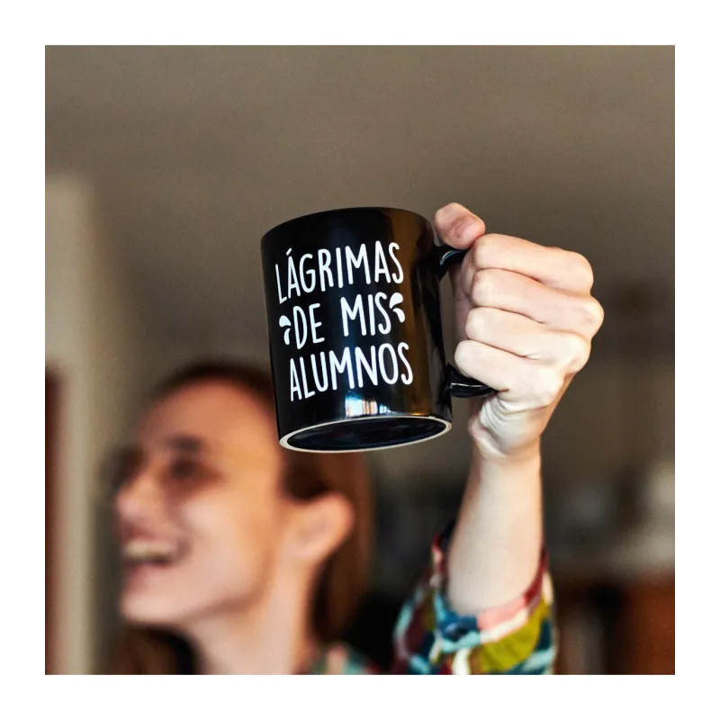 Persona levantando en alto la taza negra mostrando el texto blanco "Lágrimas de mis alumnos" - Happyideas