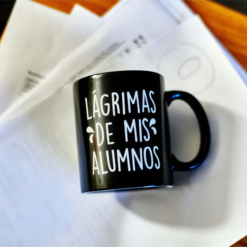 Taza negra de cerámica con texto blanco "Lágrimas de mis alumnos" apoyada sobre papeles y documentos en una mesa - Happyideas