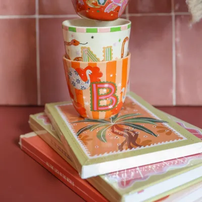 Taza Letra B, portada del producto, de cerámica con estampado floral colorido, colocada sobre una pila de libros - Happyideas