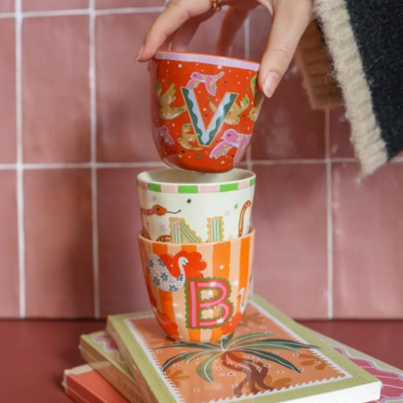 Taza Letra B apilada con otra taza monograma sobre libros, con una mano sujetándolas junto a una pared de azulejos - Happyideas