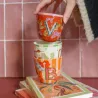 Taza Letra B apilada con otra taza monograma sobre libros, con una mano sujetándolas junto a una pared de azulejos - Happyideas
