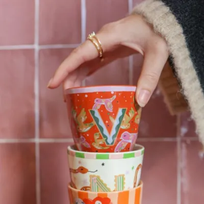 Mano sosteniendo Taza Letra V de cerámica color naranja/rojo con estampado floral, sobre fondo de azulejos rosas - Happyideas