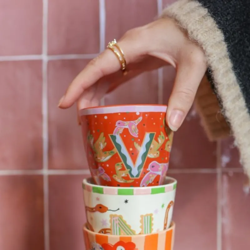 Mano sosteniendo Taza Letra V de cerámica color naranja/rojo con estampado floral, sobre fondo de azulejos rosas - Happyideas