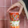 Mano sosteniendo Taza Letra V de cerámica color naranja/rojo con estampado floral, sobre fondo de azulejos rosas - Happyideas