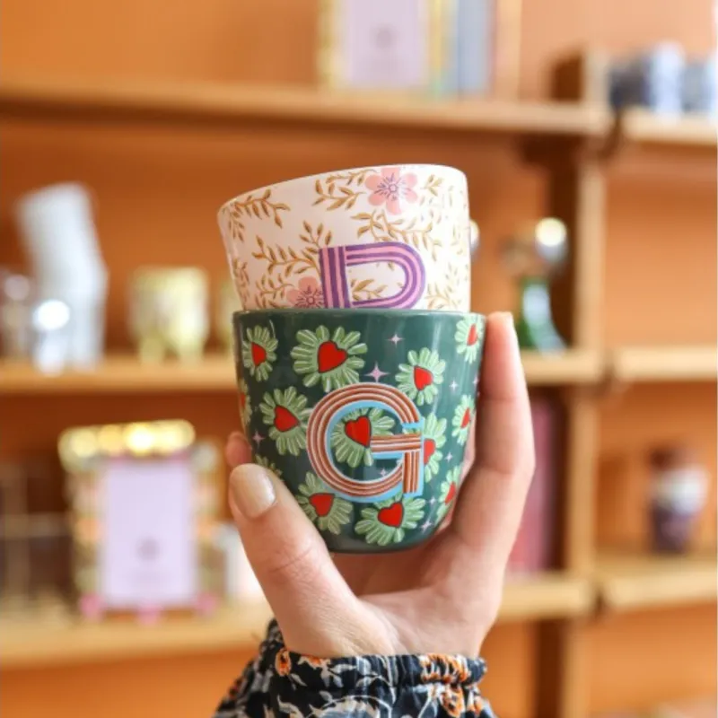 Taza Letra R sostenida en la mano, con estampado floral rosa y verde, frente a una estantería de madera - Happyideas