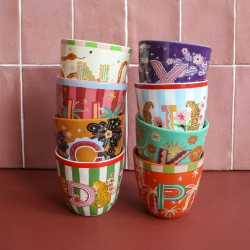 Taza Letra C junto a otras tazas monograma apiladas en dos columnas de colores frente a una pared de azulejos - Happyideas